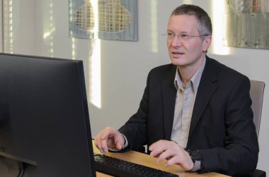 Adam bilişim ofisinde bilgisayar bölümünde çalışıyor. Gözlüklü ve ceketli orta yaşlı bir adam. Önden izleyin. Ekran görünür değil. Arkasındaki duvarda güneş parlıyor..
