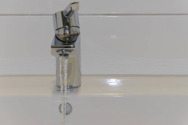 Musluktan lavaboya akan suyun yakın çekimi. Beyaz fayanslı duvar ve beyaz seramik lavabo. Arkaplan, kopyalama alanı, metin alanı