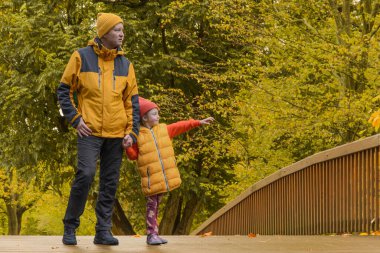 Baba ve beş yaşındaki kızı köprüdeki sonbahar parkında. Sarı ceketli adamlar uzaktaki bir şeye bakıyorlar.