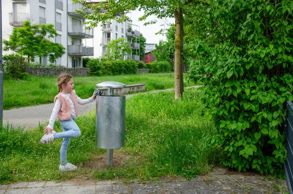Girl throws trash into a street trash can. Trash can is metal, gray. Child of five with a long blonde braid. She is wearing a cap, pink vest and blue jeans. Theres greenery all around.