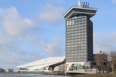 ADAM Kulesi, Amsterdam 'daki Lj Nehri' ne bakan eğimli cam panelleri olan modern bir gökdelendir. Ön planda rıhtımlı tekneleri ve açık oturaklı restoranları olan bir iskele var..