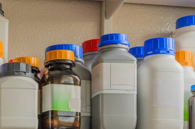 Etiketli çeşitli kimyasal şişelerle dolu bir raf. Etiketlerde tehlikeli semboller de dahil olmak üzere kimyasallar hakkında bilgi var..