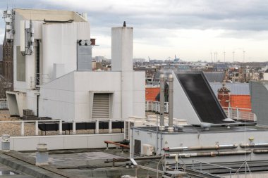 Endüstri Detaylı Çatı Manzarası. Ön planda, perçinli metal bir baca en belirgin özelliktir. Arkasında, sıcak öğleden sonra ışığında bir şehir manzarası uzanıyor..
