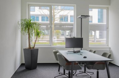 Masası ve sandalyeleri olan bir ofis tartışma odası. Arka planda büyük pencere var. Komşu bina manzaralı. Soldaki ev bitkisi yeşil palmiye. Lamba, monitör, klavye, kenetlenme istasyonu