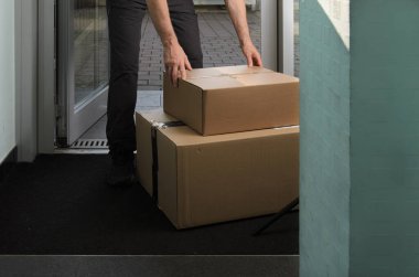 Bir kurye ön girişe iki karton paket koyar. Olay yeri teslimat anını yakalıyor, yerleşim veya ticari ortamda büyük paketlerin ele alınışını vurguluyor..