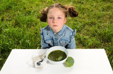 Üzgün bir çocuk bir kase brokoli çorbasına oturur. Çocuk altı yaşında mavi gözlü bir kız. Yeşil bir alana karşı beyaz bir masada oturuyor..