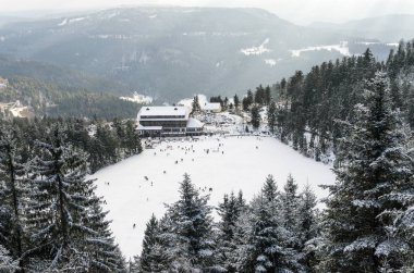 Donmuş bir göl karla kaplı ağaçlar ve uzak dağlarla çevrili ve kenarları büyük bir binayla çevrili. Birçok insan buzda görünür, kış etkinliklerinin tadını çıkarırlar..
