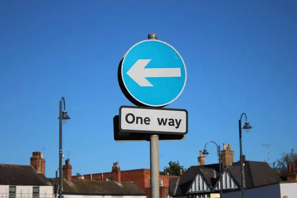 UK circular one way sign attached to a pole, blue sky and houses in background blurred