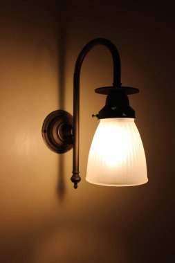 Krem rengi bir duvara tutturulmuş bronz renkli abajur, kapalı alanda aydınlatmalı.
