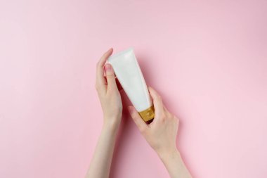Pastel pembe arka planda beyaz bir tüp tutan bir kadın.
