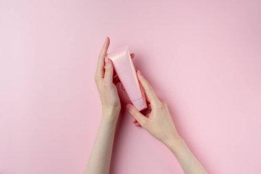 Kadının elleri pembe arka planda pembe bir tüp tutuyor.