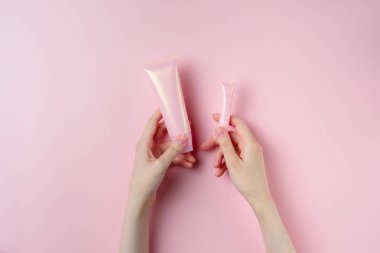 Bir kadının eli, pastel pembe arka planda iki pembe tüp tutuyor.