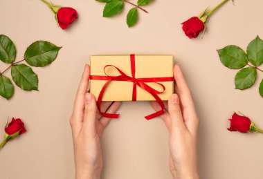 Kadın eller yukarıdan bej arka planda gül çerçevesinde bir hediye kutusu tutuyor. Düz yatış, üst görünüm.