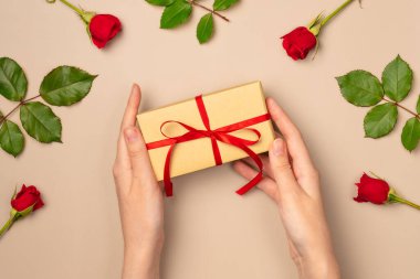 Kadın eller yukarıdan bej arka planda gül çerçevesinde bir hediye kutusu tutuyor. Düz yatış, üst görünüm.