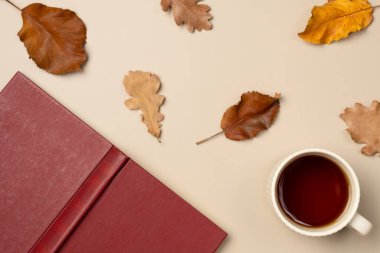 Bej arka planda kitap, yaprak ve bir fincan çay ile sonbahar kompozisyonu. Boş zaman konsepti, kitap okumak. Düz yatış, üst görünüm