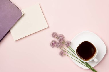 Kitap, boş kâğıt, pembe arka planda çiçekli bir fincan kahve. Tebrik kartı. Mesaj için yer var. Düz yatış, üst görünüm.