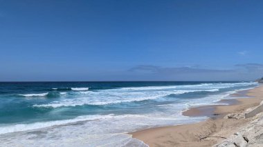 Gökyüzü açık mavi gökyüzü altında kumlu sahile vuran gök mavisi dalgalar, sakin sahil manzarası
