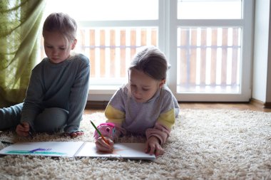 İki küçük tatlı kız, büyük bir pencerenin yanında yerde yatan bir albümde resim çiziyor..