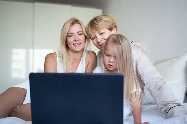 little girl with mom and grandmother are sitting on the bed in front of a laptop.