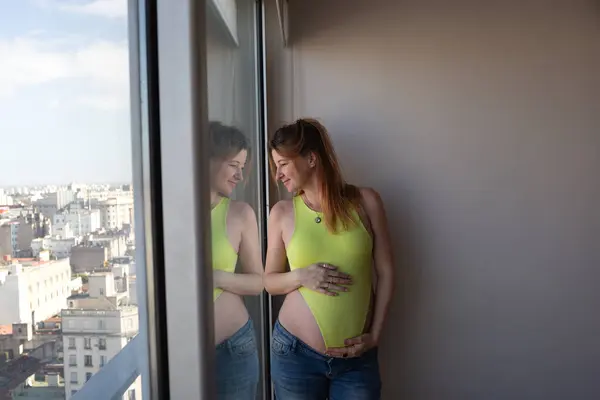 stock image A pregnant woman in a yellow bodysuit smiles, reflecting by the window, anticipating her future.