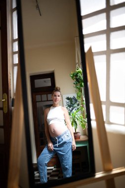Aynadan yansıyan ve doğal ışıkla çevrili gündelik kıyafetlerle hamile bir kadın..