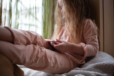 Küçük tatlı kız pencere kenarında oturmuş oyun konsolu oynuyor..