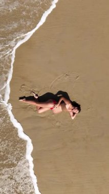 Güzel 4K dikey drone, okyanus dalgaları yavaşça kıyıya yaklaşırken sıcak kumda yatan hamile bir kadının görüntüsünü yakınlaştırıyor. Yukardan çekilen huzurlu bir annelik anı, sükunet, doğal güzellik ve zamansız uyum dolu..