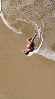 Güzel 4K dikey drone, okyanus dalgaları yavaşça kıyıya yaklaşırken sıcak kumda yatan hamile bir kadının görüntüsünü yakınlaştırıyor. Yukardan çekilen huzurlu bir annelik anı, sükunet, doğal güzellik ve zamansız uyum dolu..