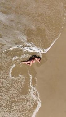 Güzel 4K dikey drone, okyanus dalgaları yavaşça kıyıya yaklaşırken sıcak kumda yatan hamile bir kadının görüntüsünü yakınlaştırıyor. Yukardan çekilen huzurlu bir annelik anı, sükunet, doğal güzellik ve zamansız uyum dolu..