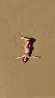 Güzel 4K dikey drone, okyanus dalgaları yavaşça kıyıya yaklaşırken sıcak kumda yatan hamile bir kadının görüntüsünü yakınlaştırıyor. Yukardan çekilen huzurlu bir annelik anı, sükunet, doğal güzellik ve zamansız uyum dolu..