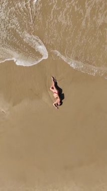 Güzel 4K dikey drone, okyanus dalgaları yavaşça kıyıya yaklaşırken sıcak kumda yatan hamile bir kadının görüntüsünü yakınlaştırıyor. Yukardan çekilen huzurlu bir annelik anı, sükunet, doğal güzellik ve zamansız uyum dolu..