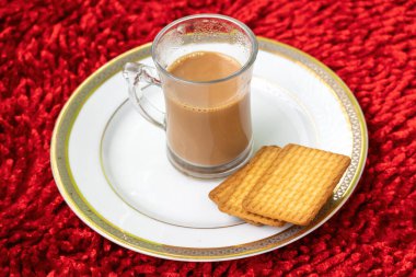 Bardakta bir fincan sıcak süt çayı ve kırmızı halıda beyaz bir tabakta biraz kurabiye..