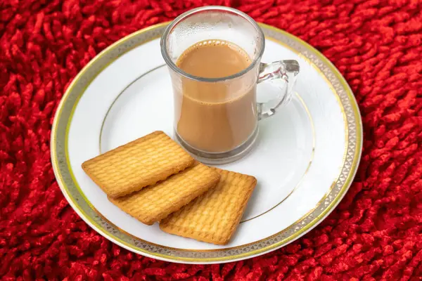 Kırmızı halı desenli arka planda beyaz bir tabakta bisküviler olan cam bir fincanda bir fincan sıcak süt çayı..