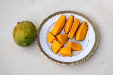 Ahşap zemin üzerinde beyaz bir tabakta soyulmuş mango dilimleri, yanında olgun bir mango.