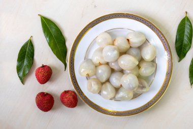 Hafif ahşap yüzeyde beyaz bir tabakta taze soyulmuş lychees. Yanında biraz litchi ve yeşil yapraklar olsun. Üst görünüm.