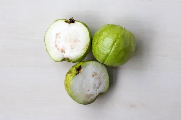 stock image One whole fresh guava fruit and one cut into two slices, displayed on light wooden surface. Top view.