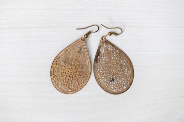 Lüks çiçek desenleri ve metal kanca tokaları olan bir çift antika küpenin üst görüntüsü, açık ahşap bir arka planda sergilenmektedir..