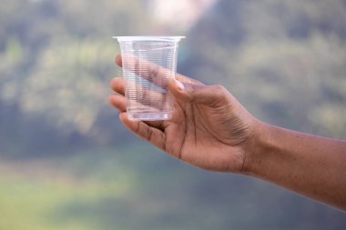 Bir kadın eli, plastik kirlilik ve çevre sorunları kavramını gösteren bulanık bir arka plan karşısında boş bir plastik bardak tutuyor..