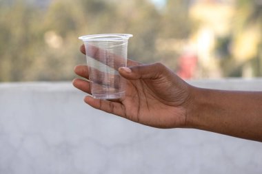 Bir kadın eli, plastik kirlilik ve çevre sorunları kavramını gösteren bulanık bir arka plana karşı boş bir plastik bardak tutuyor..