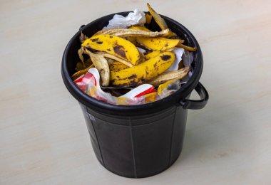 Ev çöpü ve atıklarla dolu siyah plastik bir kova. Görünür muz kabukları, kağıt, mendil, politen torbaları..
