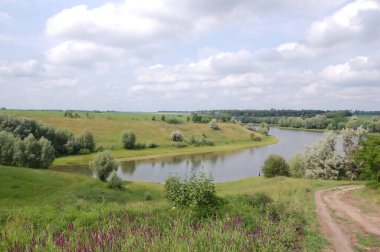 Yazın kırsal kesimde Manzaralı Nehir Eğrisi. Yeşil tepeler boyunca kıvrılan sakin bir nehrin ve parçalı bulutlu bir gökyüzünün altında açık çayırların güzel panoramik manzarası. Manzarada yemyeşil otlar serpilmiştir.