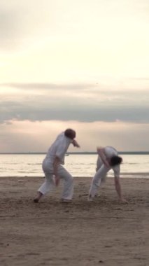 Beyaz elbiseli iki adam sahilde capoeira çalışıyor. Dikey video