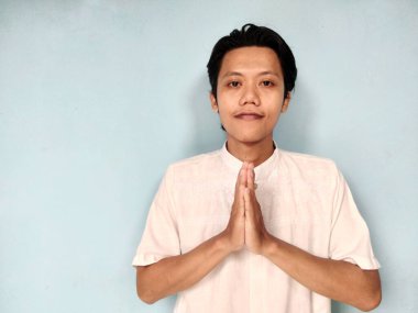 Beyaz gömlek giyen genç Asyalı adam yüzünde kocaman bir gülümsemeyle ona selam veriyor. Gri arka planda izole. Bayram Mübarek, Bayram Fitr, Kurban Bayramı selamlaması