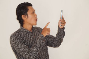 Siyah tişörtlü genç Asyalı adam şaşırdı ve beyaz arka planda izole akıllı telefona işaret etti.