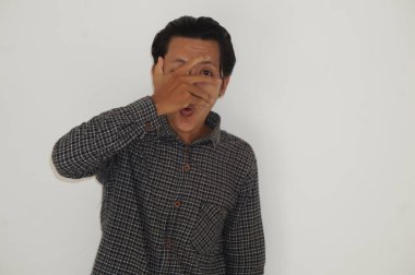 Siyah gömlekli Asyalı genç adam. Yüzünü ve gözlerini elleriyle kapatıyor, parmaklarının arasından beyaz bir arka plana bakıyor.