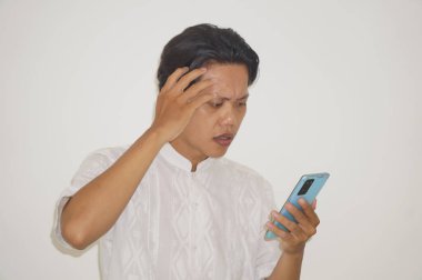 Beyaz tişörtlü genç Asyalı adam beyaz arka planda izole edilmiş akıllı telefona bakarken şaşkın hissediyordu..