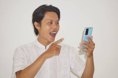 Beyaz tişörtlü genç Asyalı adam gülümsüyor ve beyaz arka planda izole akıllı telefonu işaret ediyor.