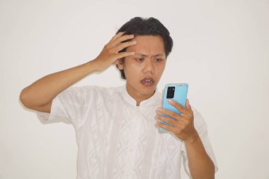 Beyaz tişörtlü genç Asyalı adam beyaz arka planda izole edilmiş akıllı telefona bakarken üzgün hissediyor..