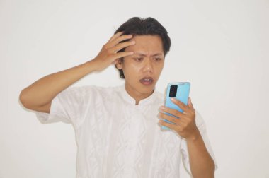 Beyaz tişörtlü genç Asyalı adam beyaz arka planda izole edilmiş akıllı telefona bakarken üzgün hissediyor..