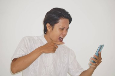 Beyaz tişörtlü genç Asyalı kızgın ve beyaz arka planda izole edilmiş akıllı telefonu işaret ediyor.